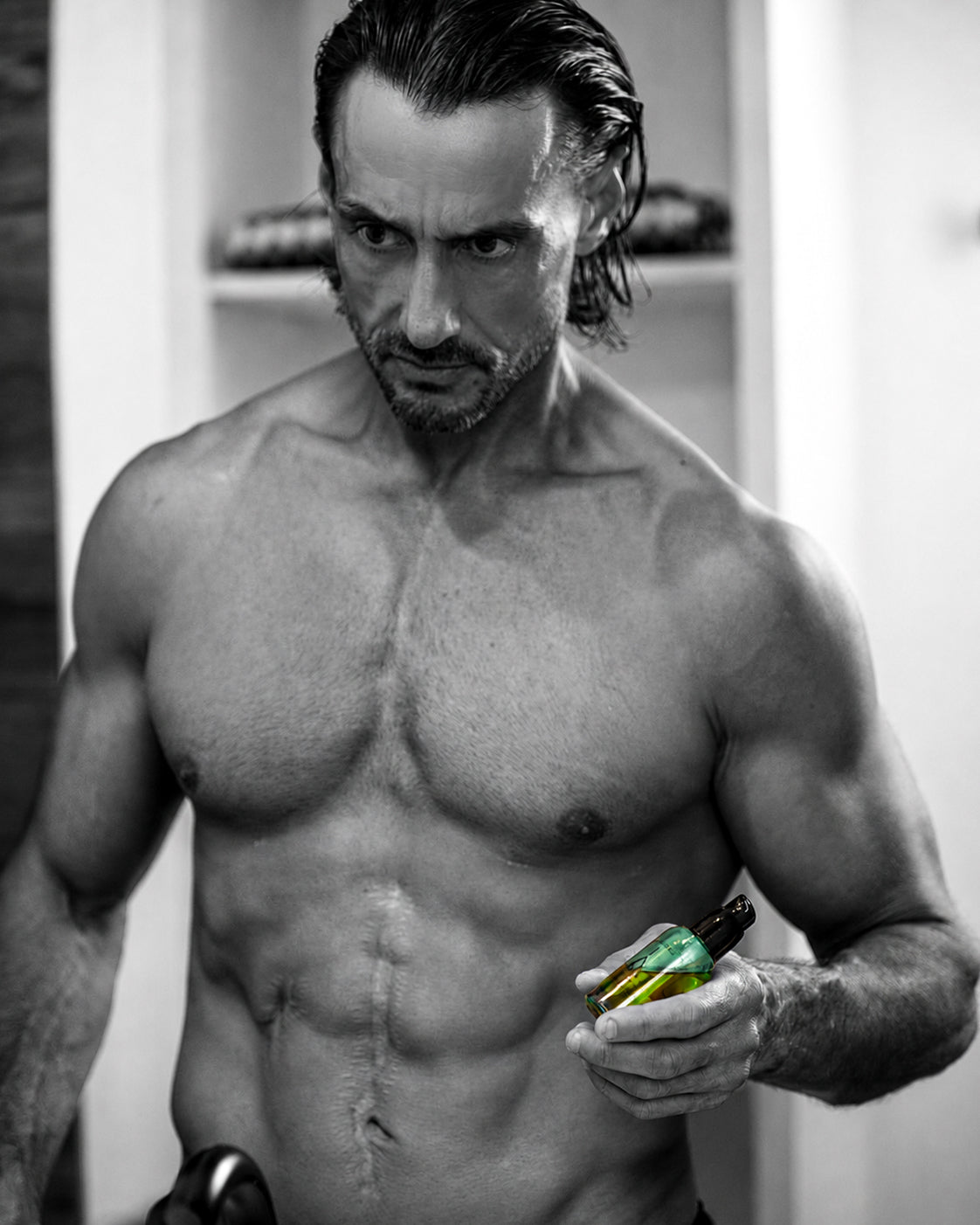A shirtless, muscular man with wet hair holds a green bottle in a bathroom setting, shown in black and white with a selective color treatment on the bottle.