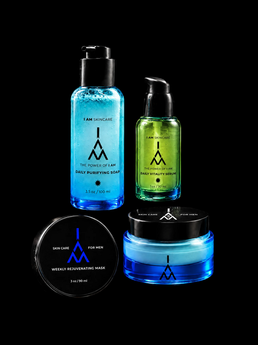 Four skincare products from I AM SKINCARE arranged on dark draped fabric and rocks: a tall blue daily purifying soap bottle, a green daily vitality serum bottle, a round blue weekly rejuvenating mask jar, and a black lid labeled weekly rejuvenating mask for men. The products feature a stylized blue 'I AM' logo.