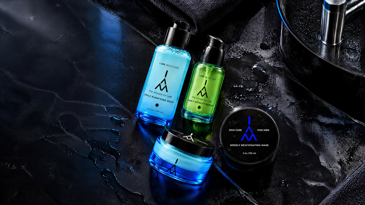 Four grooming products from Men & Men on a dark, wet surface: two blue bottles, one blue jar, and one black tin with a blue logo. The products are arranged with dramatic lighting and water droplets.